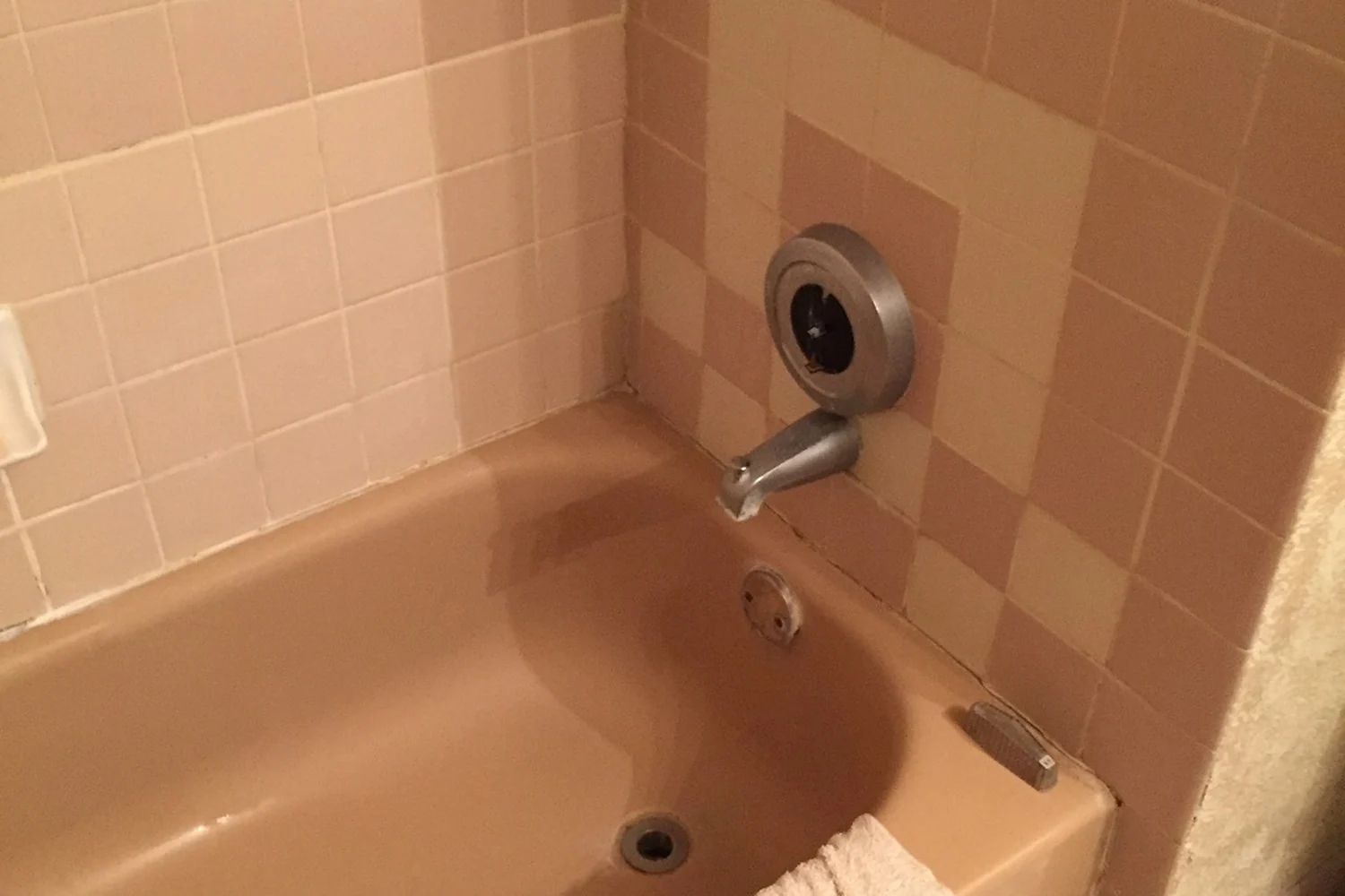 Holland bathroom tub before renovation, with a tan fiberglass tub, mismatched dated tan and pink wall tile, an aged chrome spout, and cracked grout