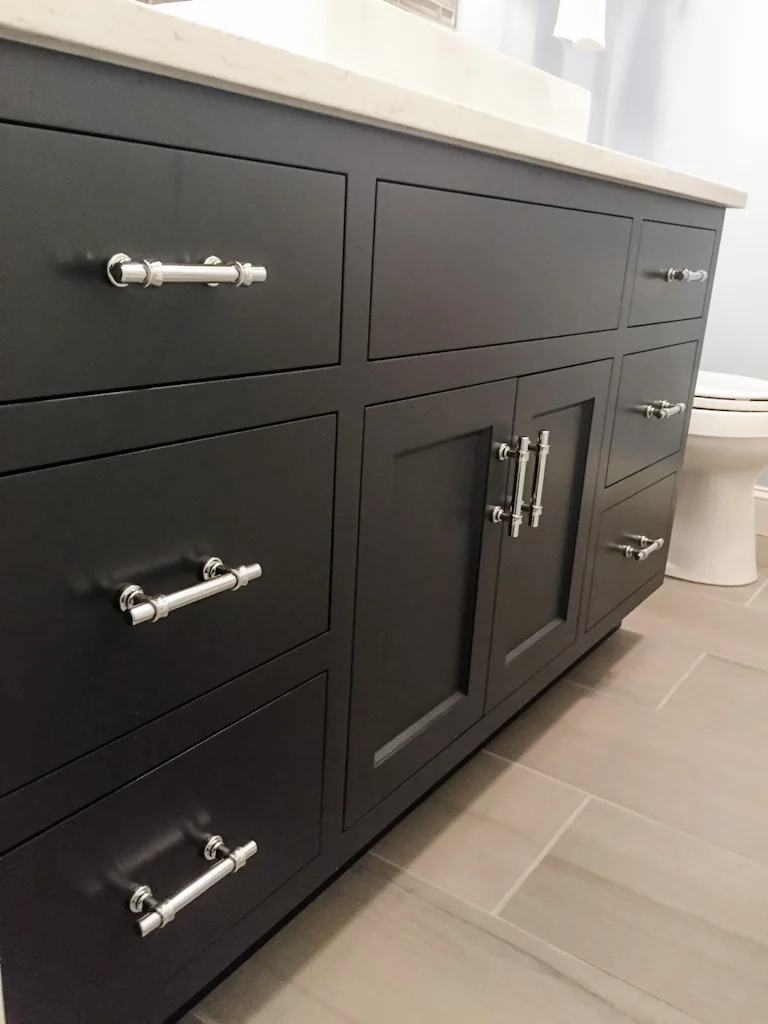 Angled view of the custom navy Nina Green Signature Series vanity cabinet in the Holland bathroom, showing the shaker drawer and door fronts, polished chrome bar pulls, and white quartz top above wood-look porcelain floor tile