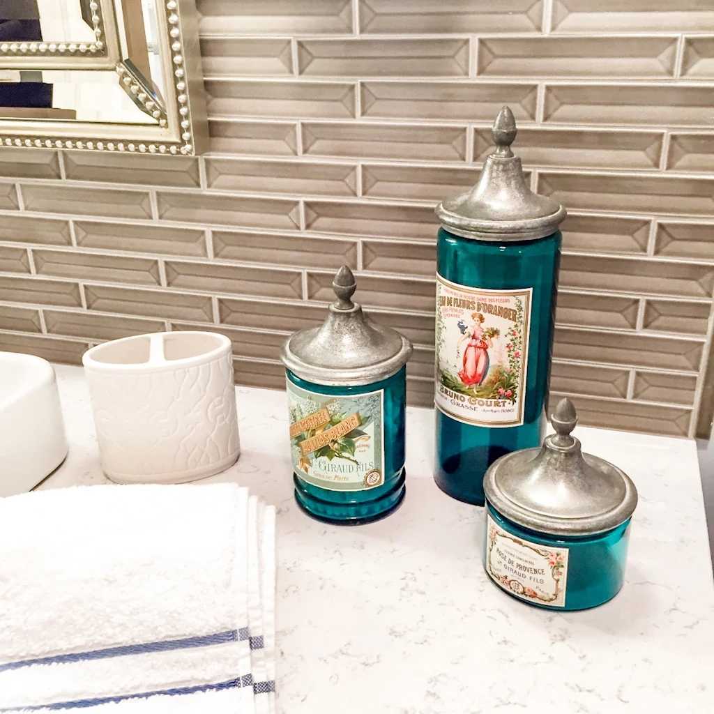 Teal apothecary jars with pewter lids and vintage French labels styled on the white quartz vanity top against the linear glass tile accent wall in the Holland contemporary bathroom