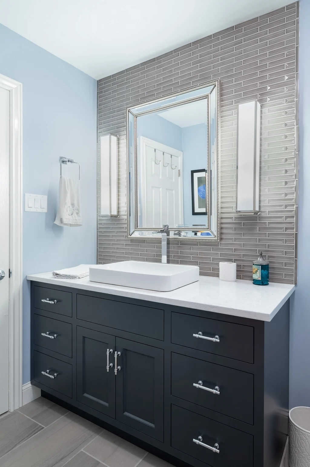Custom navy Nina Green Signature Series vanity in the Holland contemporary bathroom, with a white quartz countertop, rectangular vessel sink, polished chrome waterfall faucet, beaded silver mirror, and a linear glass tile accent wall against soft blue walls