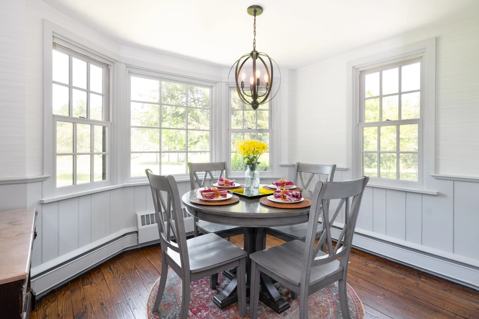 Richboro eat-in breakfast area after renovation, with a bright white bay window, soft gray board-and-batten wainscoting, a gray round pedestal table with cross-back chairs, and a globe chandelier
