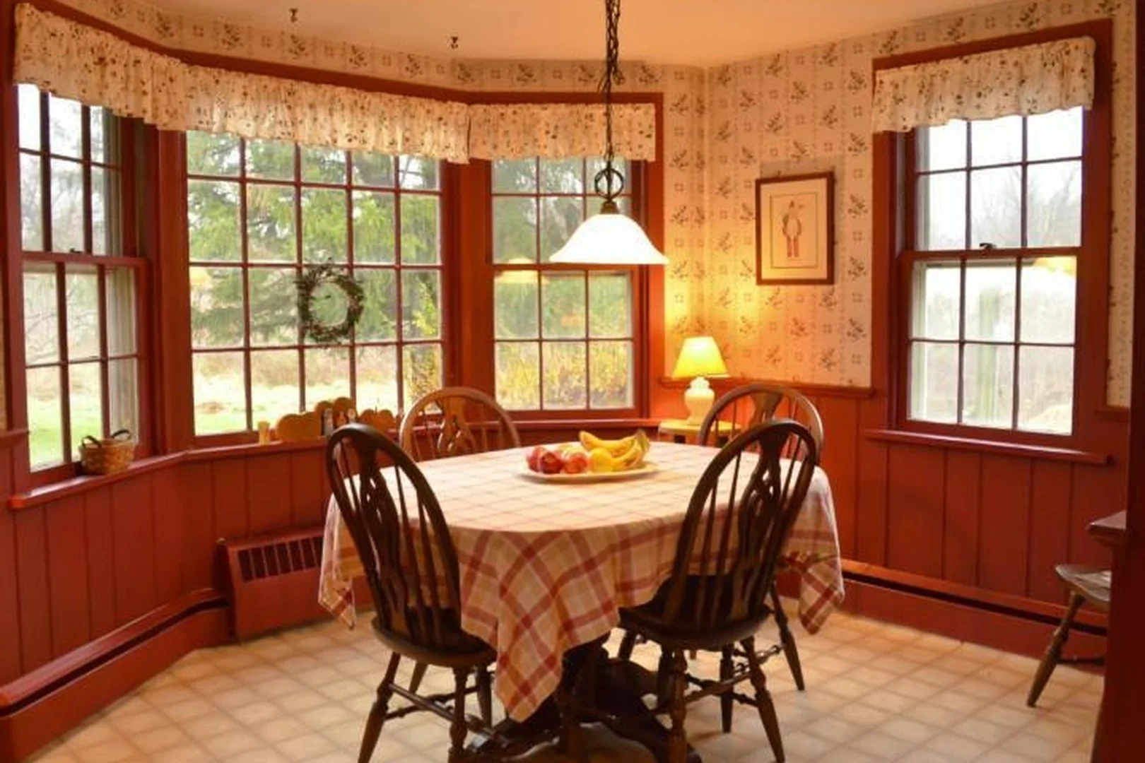 Richboro eat-in breakfast area before renovation, with a dark red bay window, floral valances and wallpaper, dark red wainscoting, and a plaid-clothed table with spindle-back chairs