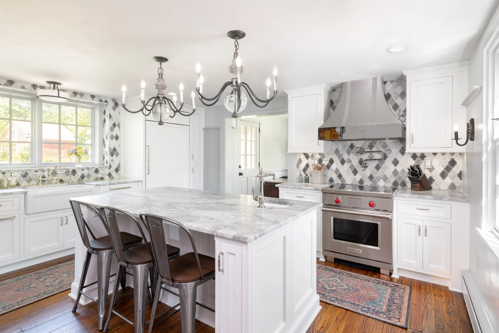 Richboro kitchen after renovation, viewed toward the back door, with white Nina Green Signature Series cabinets, a marble-topped island and industrial stools, a stainless Wolf range, arabesque marble backsplash, and wood-and-iron chandeliers