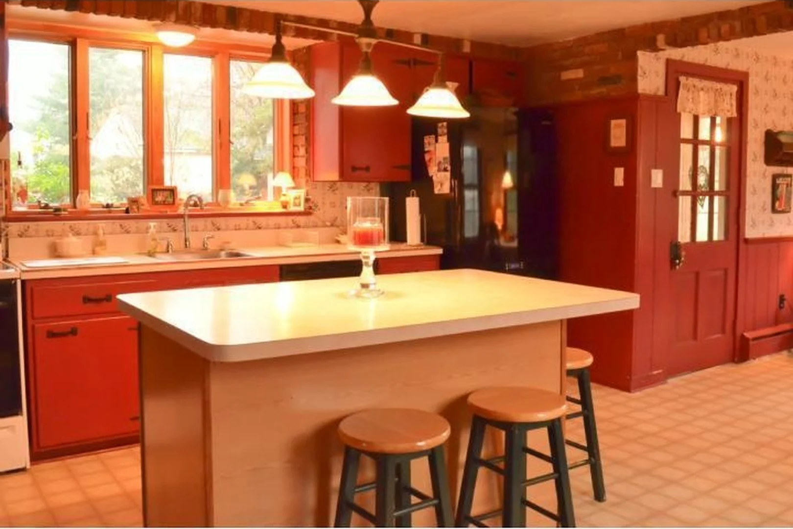 Richboro kitchen before renovation, with red painted cabinets, a maple butcher-block island and stools, dark red trim around the back door, exposed brick, and dated pendant lights