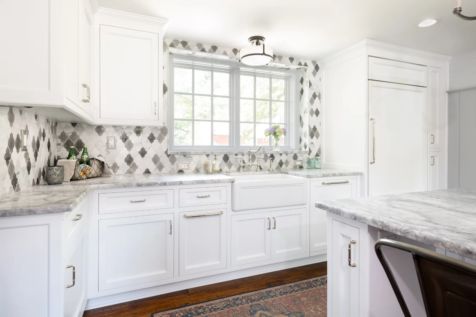 Richboro kitchen after renovation along the sink wall, with white cabinets, a farmhouse apron sink beneath the window, marble counters, an arabesque marble backsplash, and a panel-front refrigerator