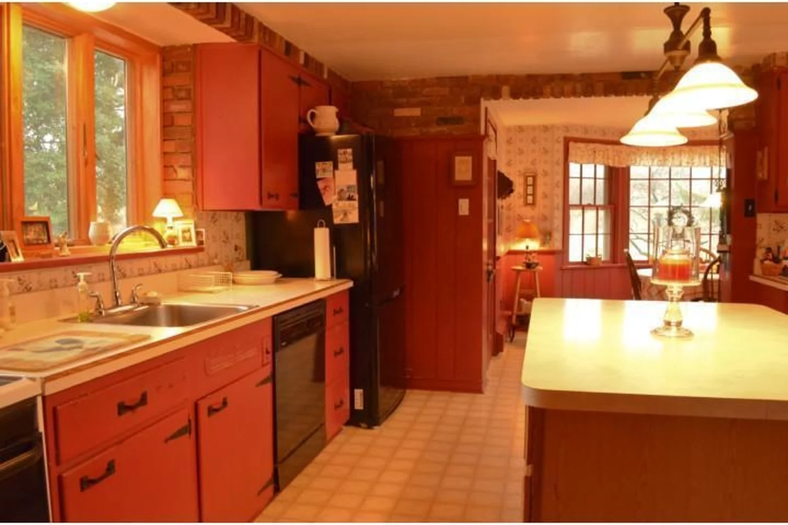 Richboro kitchen before renovation, viewed along the sink wall toward the eat-in area, with red cabinets, a black refrigerator, laminate counters, and a worn vinyl floor