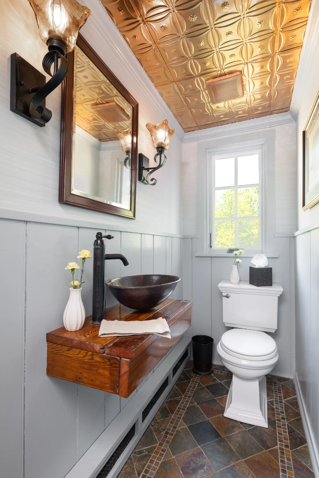 Full view of the Richboro powder room with a hammered-copper vessel sink on a reclaimed-wood shelf, gray board-and-batten wainscoting, slate tile floor, and a glowing pressed-copper tin ceiling