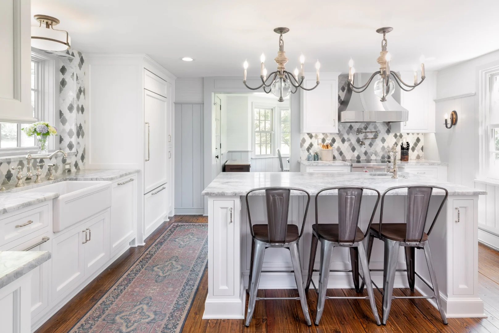 Renovated Richboro farmhouse kitchen with white Nina Green Signature Series cabinets, a marble-topped island with industrial counter stools, an arabesque marble backsplash, a stainless range, and a pair of wood-and-iron chandeliers over wide-plank wood floors
