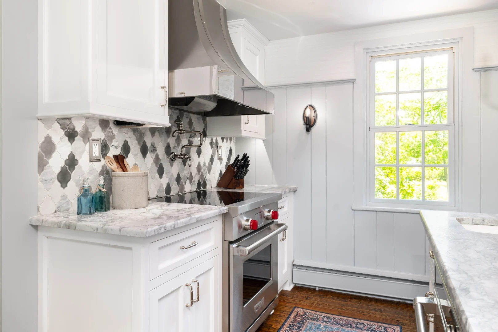 Stainless Wolf cooktop and range with a curved stainless hood and articulating pot filler against an arabesque marble backsplash in the Richboro kitchen, beside a sunny board-and-batten window wall
