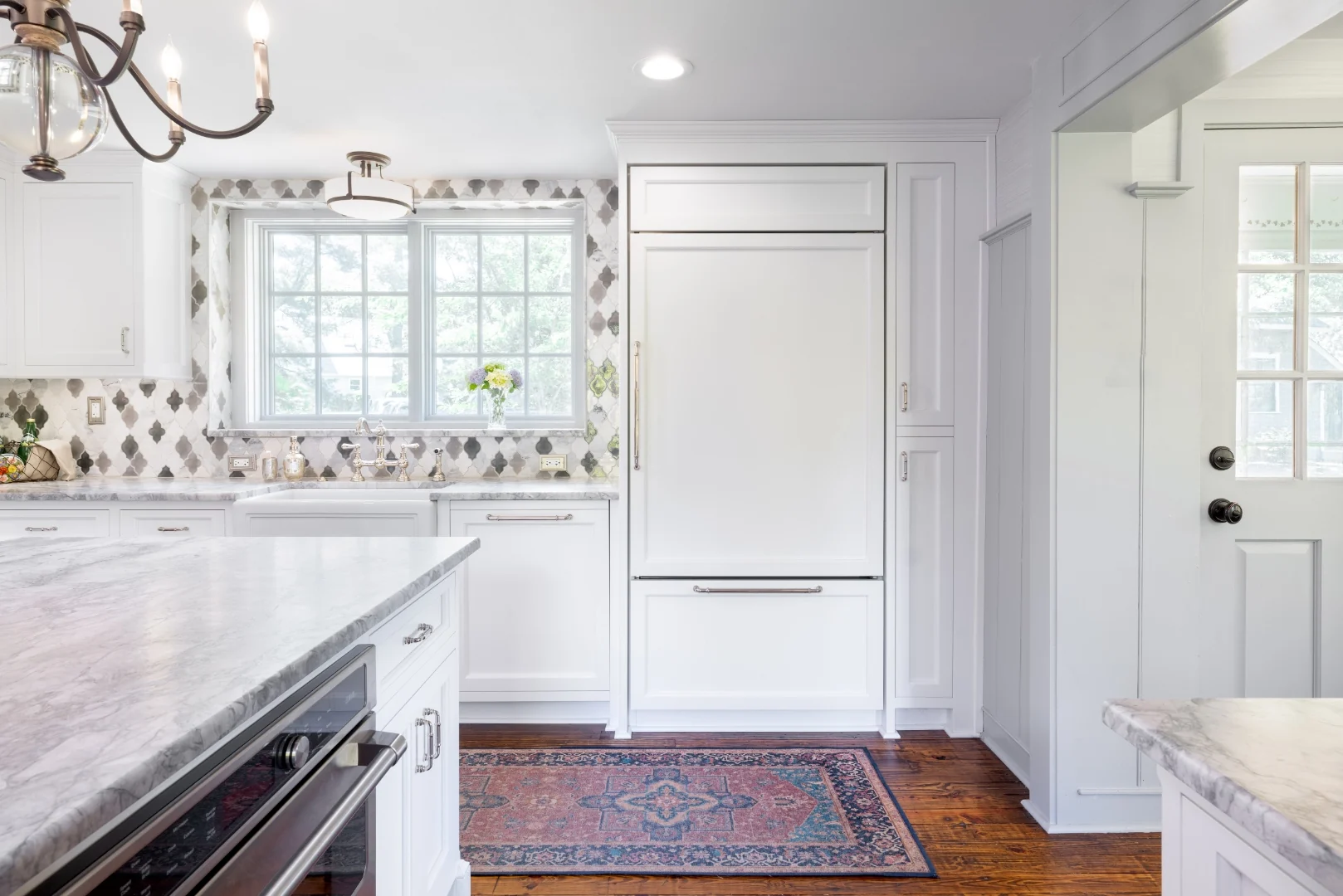 Panel-front refrigerator and tall white pantry cabinet in the Richboro kitchen, beside a built-in wall oven in the island and the back door with a wood-and-iron chandelier overhead
