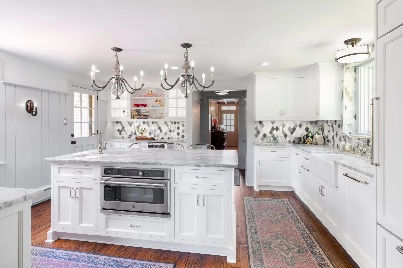 Wide view of the Richboro kitchen with a large marble-topped island housing a built-in oven, white Nina Green Signature Series cabinets, twin chandeliers, an open glass-front hutch, and a view through to the hallway