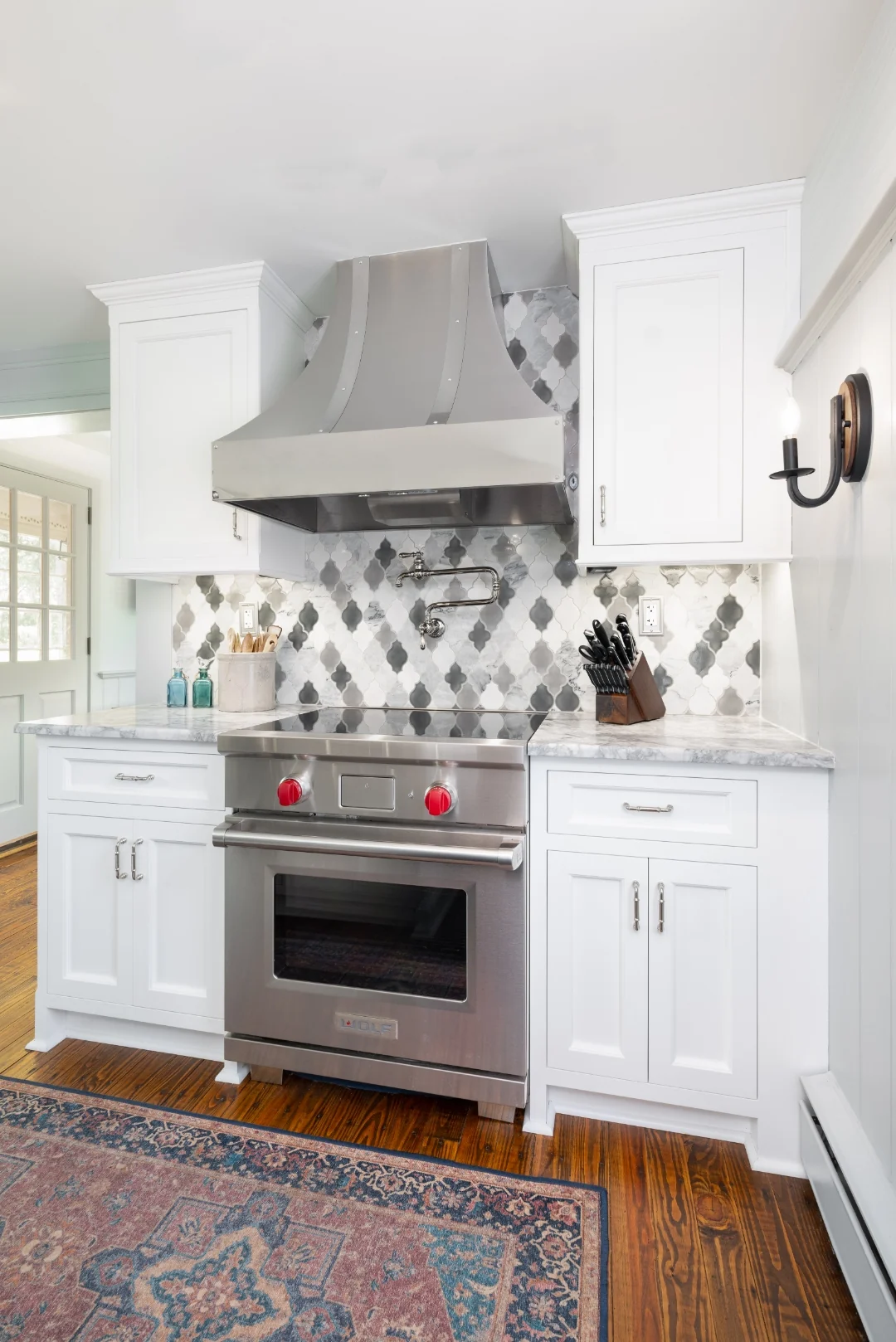 Stainless Wolf range with red knobs centered beneath a curved stainless hood and arabesque marble backsplash, flanked by white Nina Green Signature Series cabinets in the Richboro kitchen