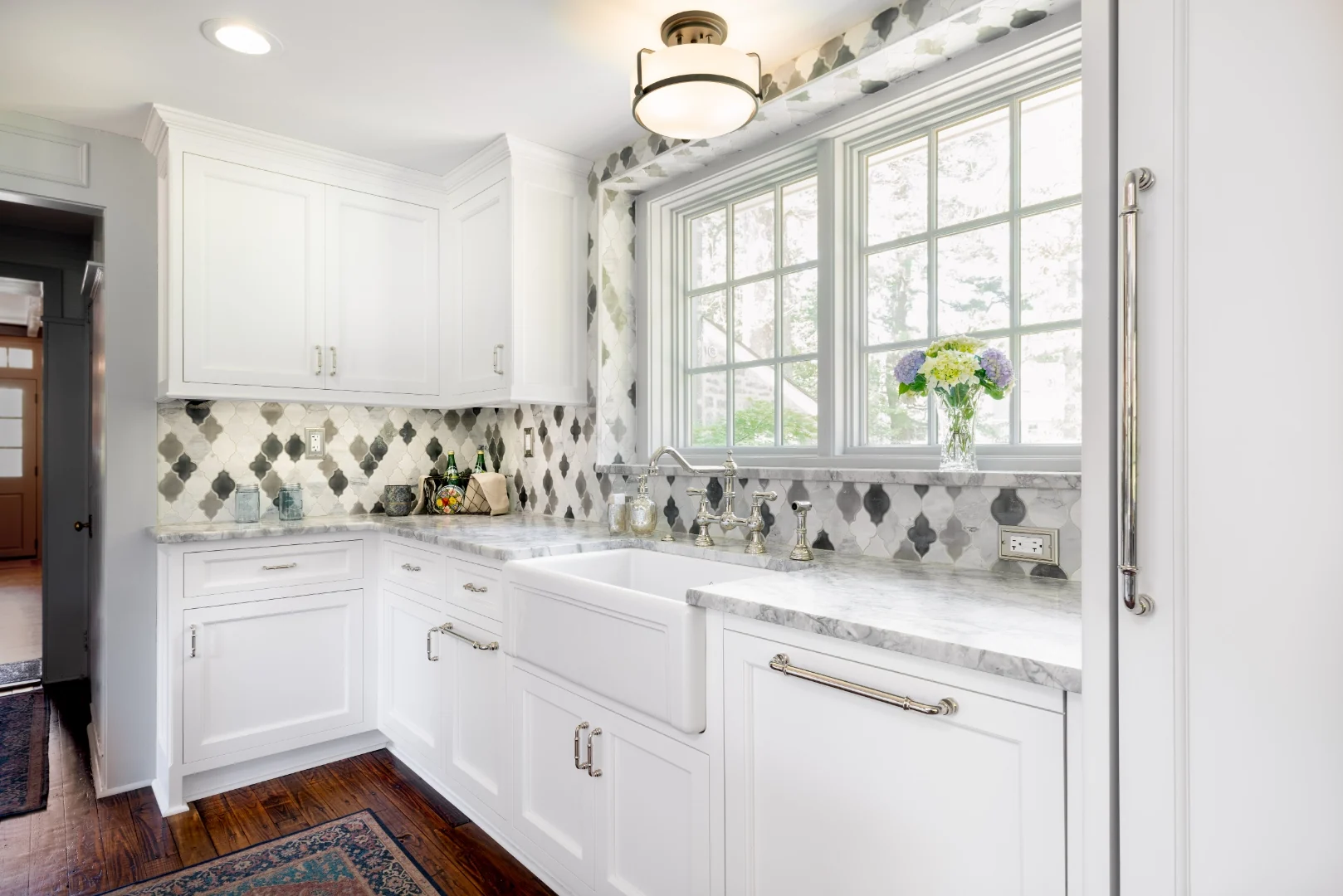 Corner of the Richboro kitchen with a farmhouse apron sink, polished-nickel bridge faucet, marble counters, arabesque marble backsplash, and a panel-front refrigerator with cup-pull hardware