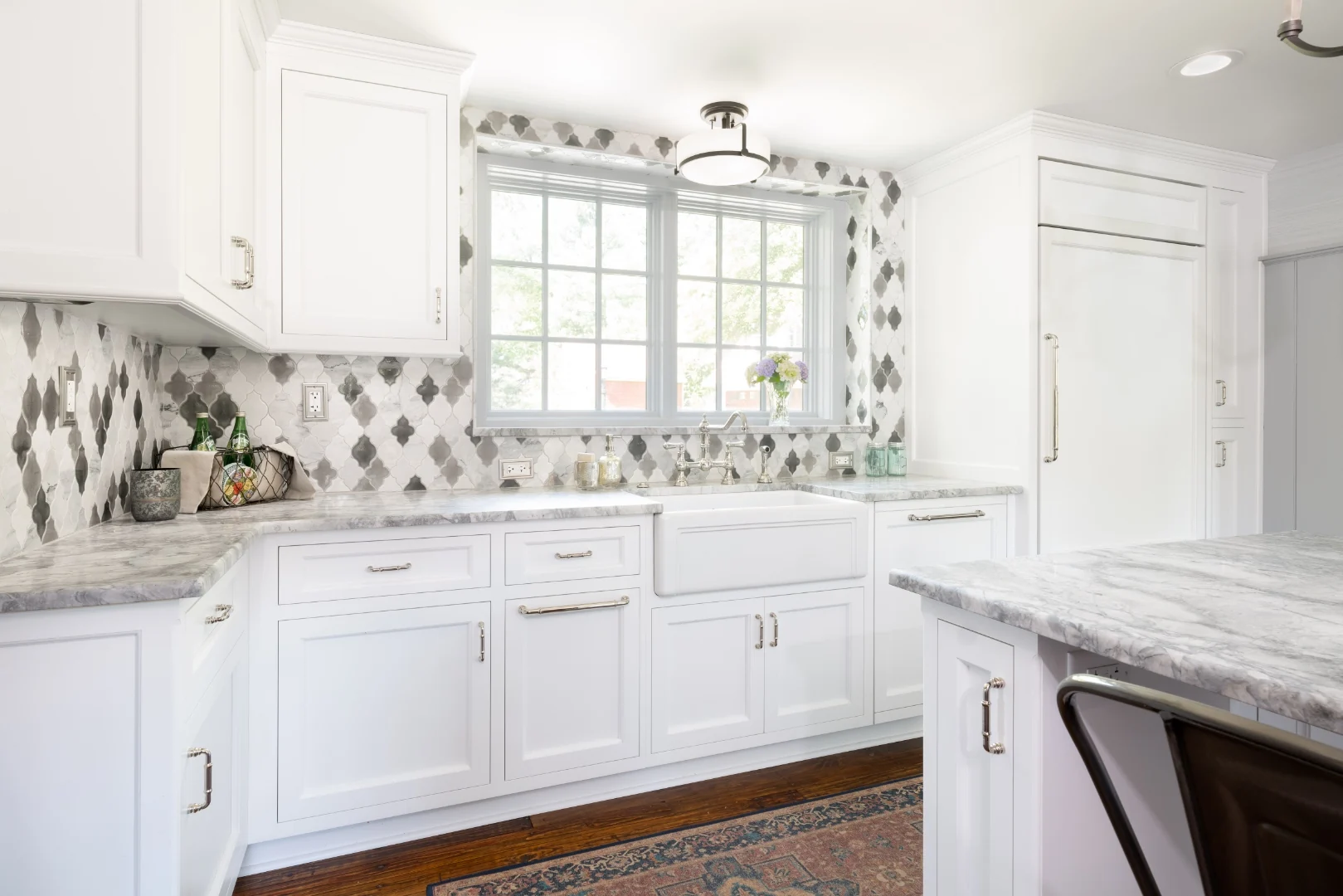 Farmhouse apron-front sink beneath a window in the Richboro kitchen, with white cabinets, polished-nickel bridge faucet, marble counters, and a gray-and-white arabesque marble backsplash