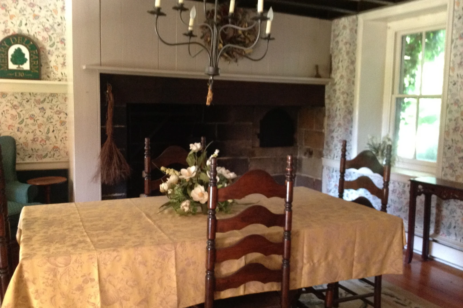 Southampton dining room before renovation, with busy floral wallpaper, dark exposed beams, a damask tablecloth on a dark wood table, and ladder-back chairs in front of the original stone fireplace