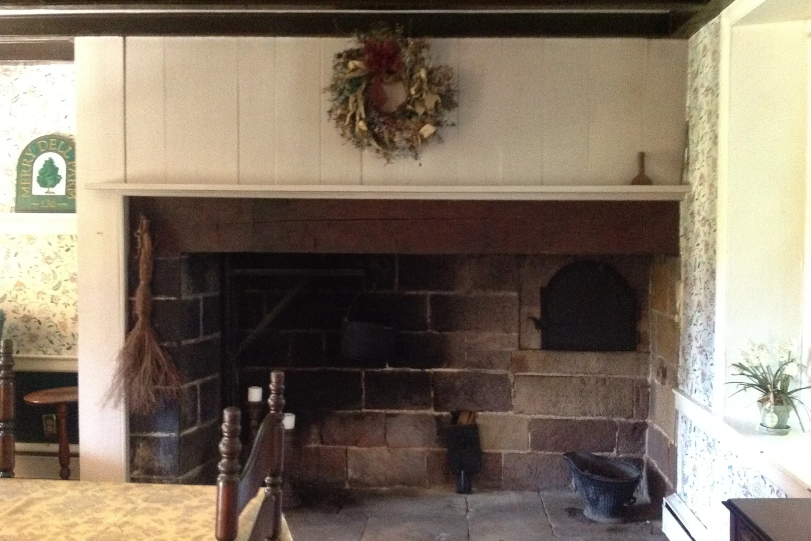 The original walk-in cooking hearth before restoration — a white-painted overmantel, dried-flower wreath, and a soot-darkened stone firebox with a beehive bake oven