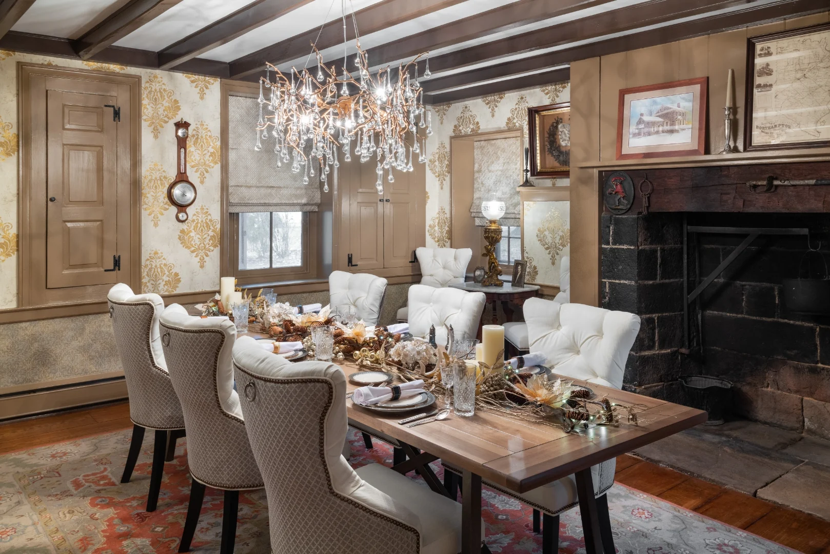 The branch-and-crystal chandelier hanging above the set dining table, framed by gold damask wallpaper and the blackened stone hearth in the Southampton dining room