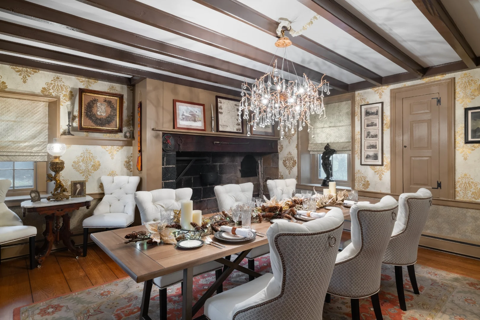 Restored Southampton historic dining room with gold metallic damask wallpaper, white tufted dining chairs, a branch-and-crystal chandelier, and the original stone walk-in cooking hearth, set with a lavish gold holiday tablescape