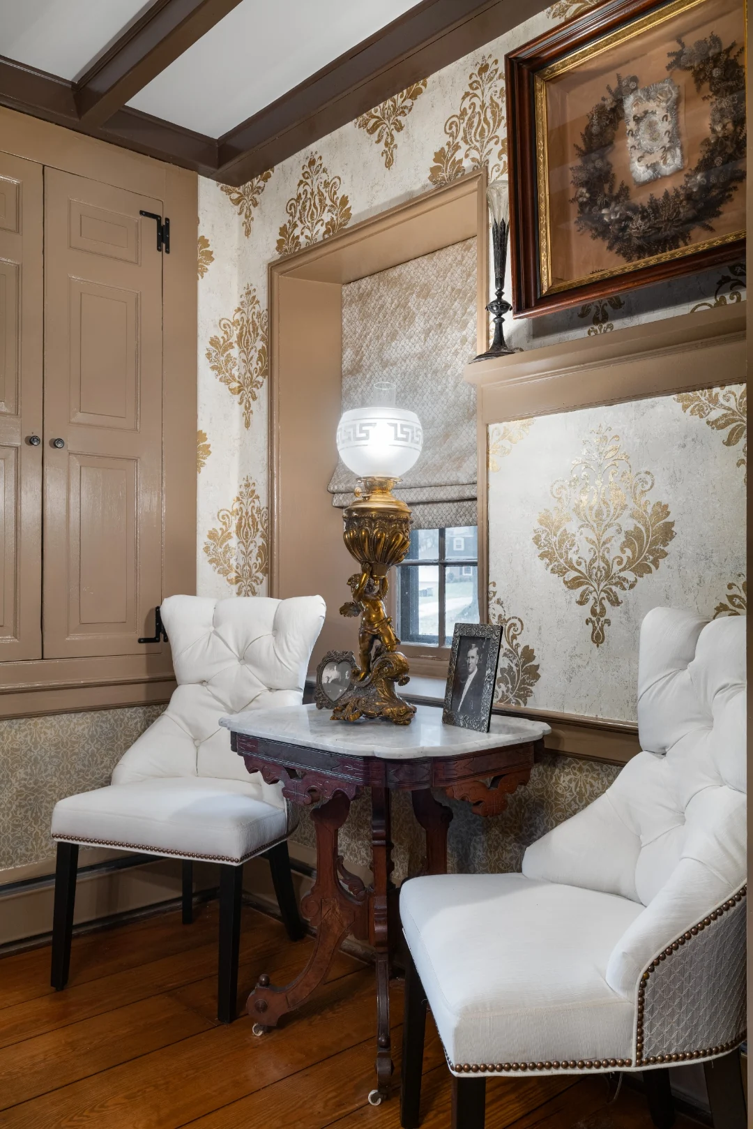 Corner vignette with a marble-topped Victorian parlor table, gilt cherub oil lamp, and white tufted chairs beneath a framed mourning wreath in the Southampton dining room