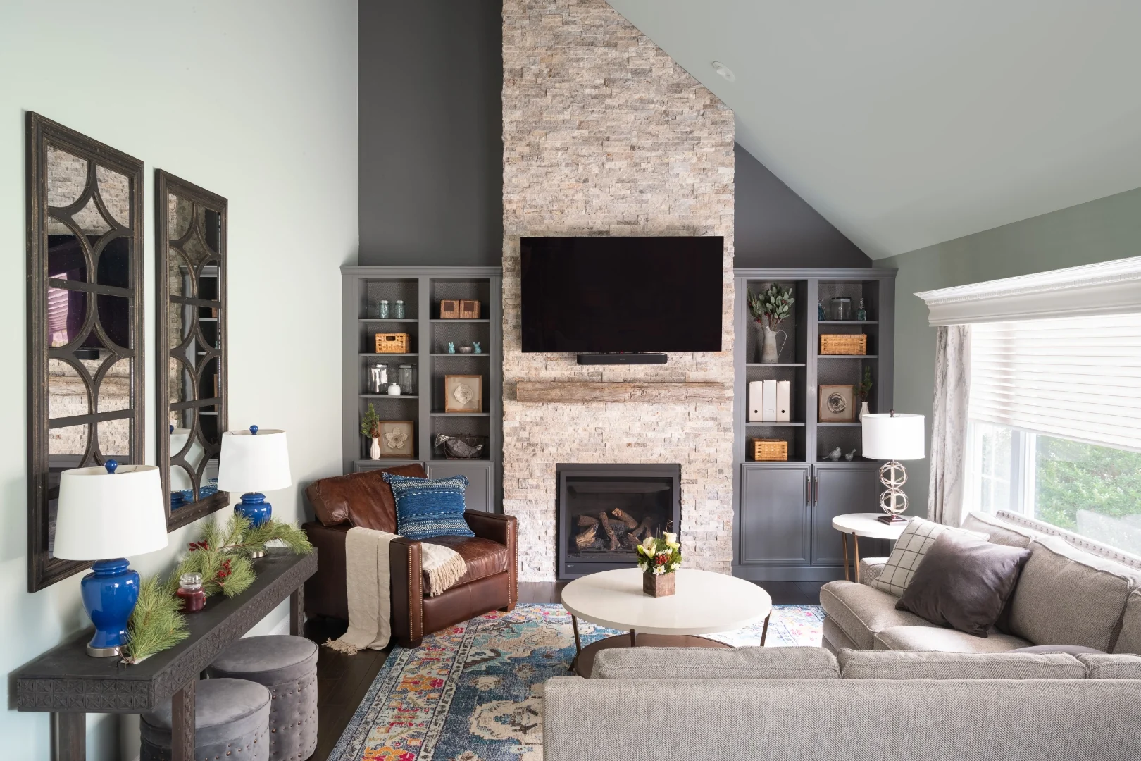 Yardley living room fireplace wall after renovation, with a dramatic floor-to-ceiling stacked stone fireplace, a reclaimed wood mantel beam, a mounted TV, custom gray built-in bookcases, and a leather club chair beneath the vaulted ceiling