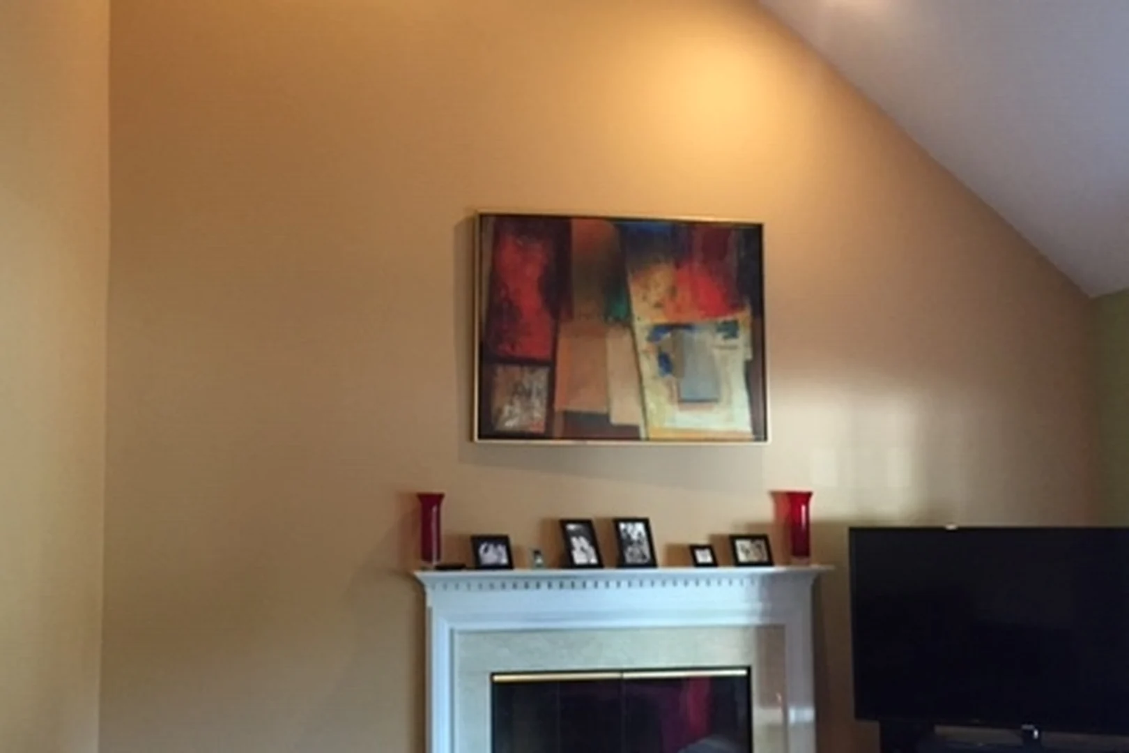 Yardley living room fireplace wall before renovation, with a small white tile fireplace, an abstract painting on a tall tan vaulted wall, framed photos on a plain mantel, and a TV on a black stand