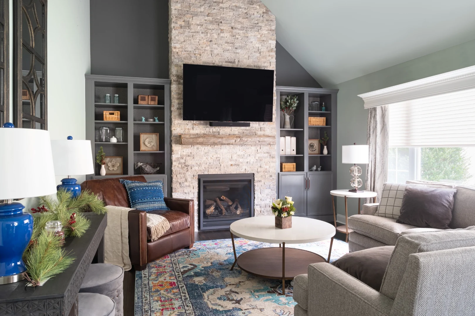Yardley living room after renovation, a bright transitional rustic space with herringbone sofas, a leather club chair, a round coffee table, a blue Persian rug, custom gray built-ins, and the stacked stone fireplace beneath the vaulted ceiling