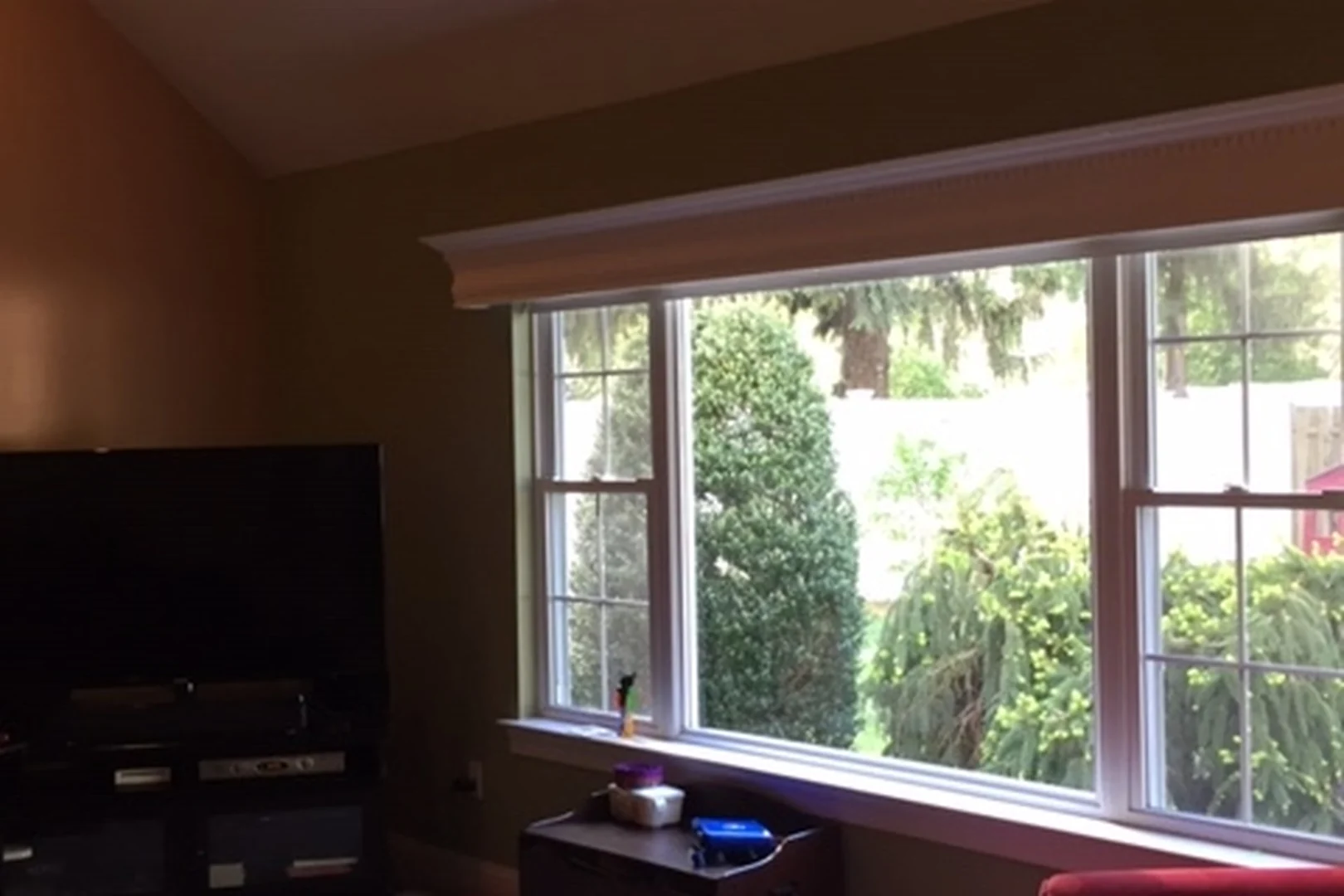 Yardley living room before renovation, viewed toward the picture windows, with dim olive walls, a TV on a black stand, a red sectional, and a cluttered window-side cabinet beneath the vaulted ceiling