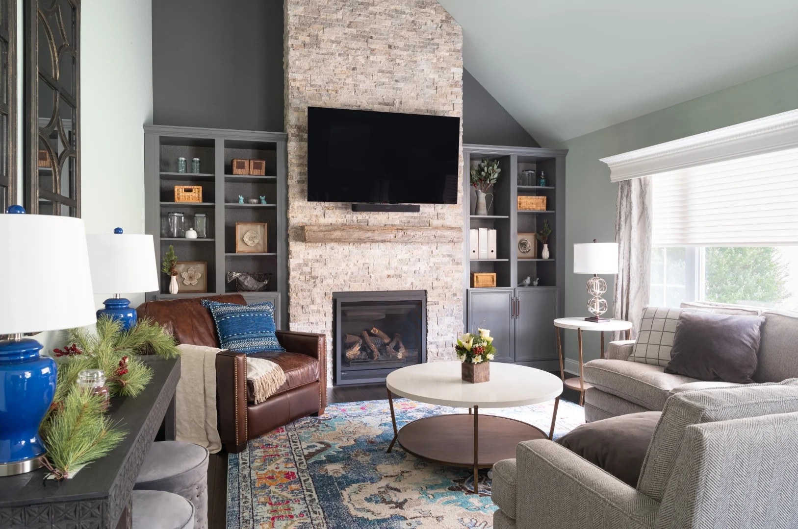 Yardley transitional rustic living room with a floor-to-ceiling stacked stone fireplace, a reclaimed wood mantel beam and mounted TV, custom gray built-in bookcases, a leather club chair, herringbone sofas, and a blue Persian rug beneath a vaulted ceiling