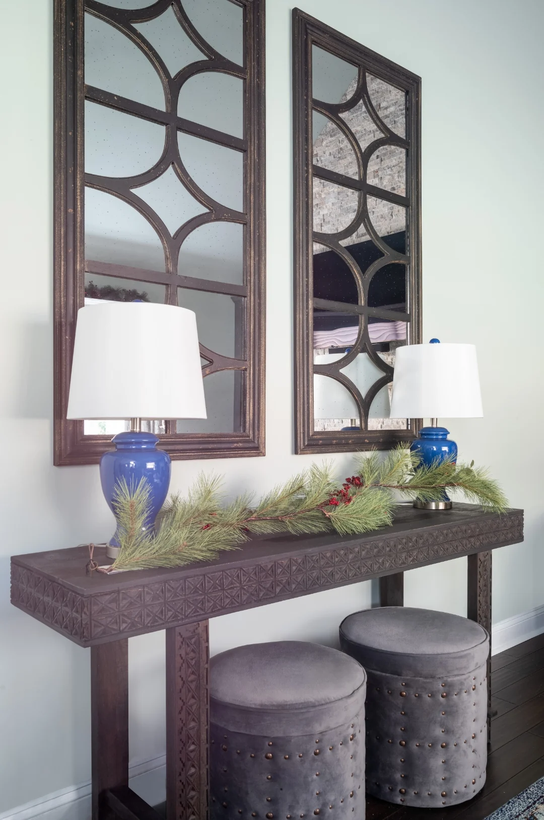 Carved dark-wood console table with twin blue ginger-jar lamps and a winter pine garland beneath a pair of arched lattice mirrors, with nailhead storage stools tucked below, in the Yardley living room