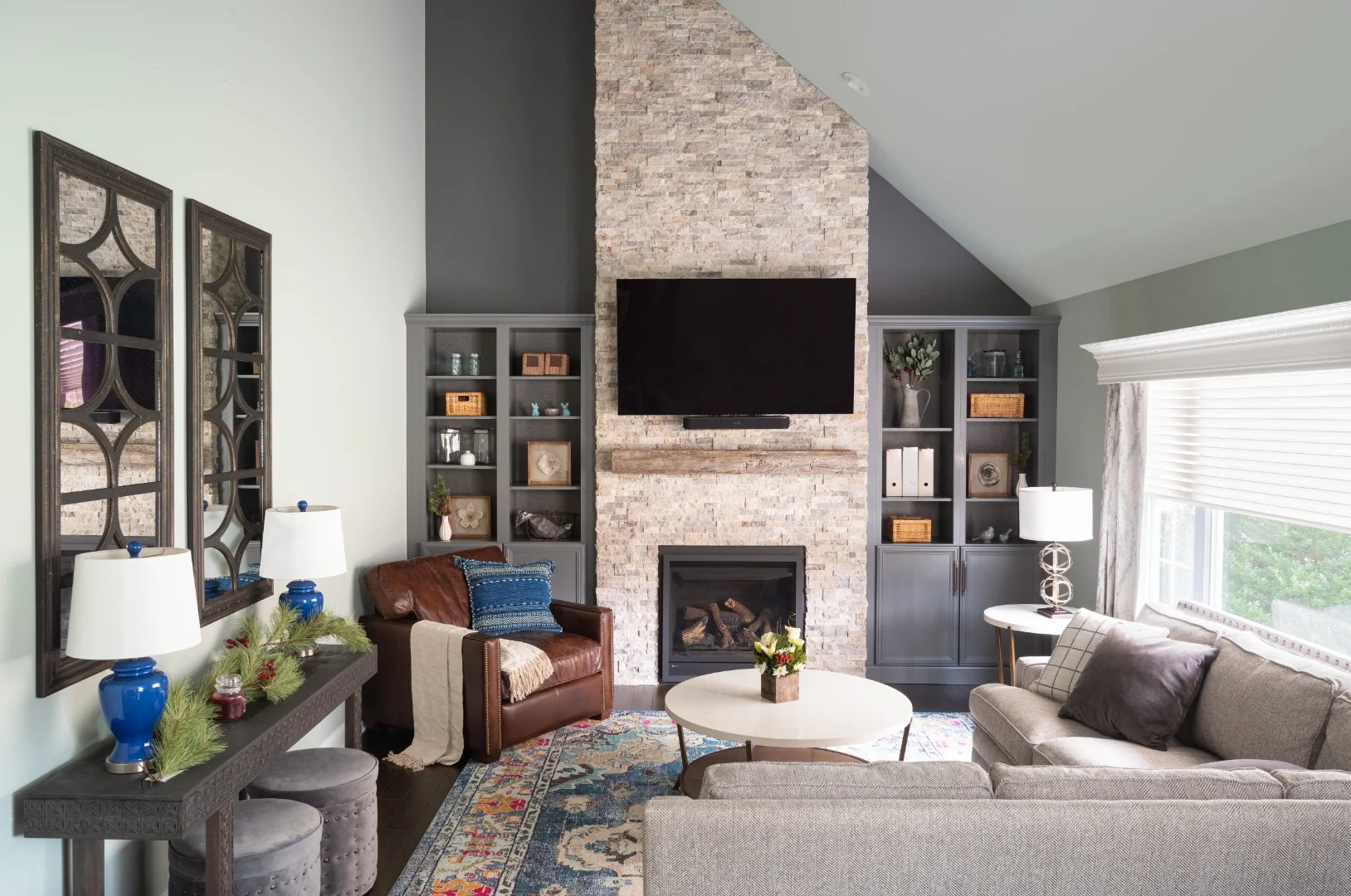 Wide view of the Yardley transitional rustic living room showing the stacked stone fireplace and reclaimed wood mantel flanked by gray built-in bookcases, a console with twin arched mirrors, herringbone sofas, and a leather club chair