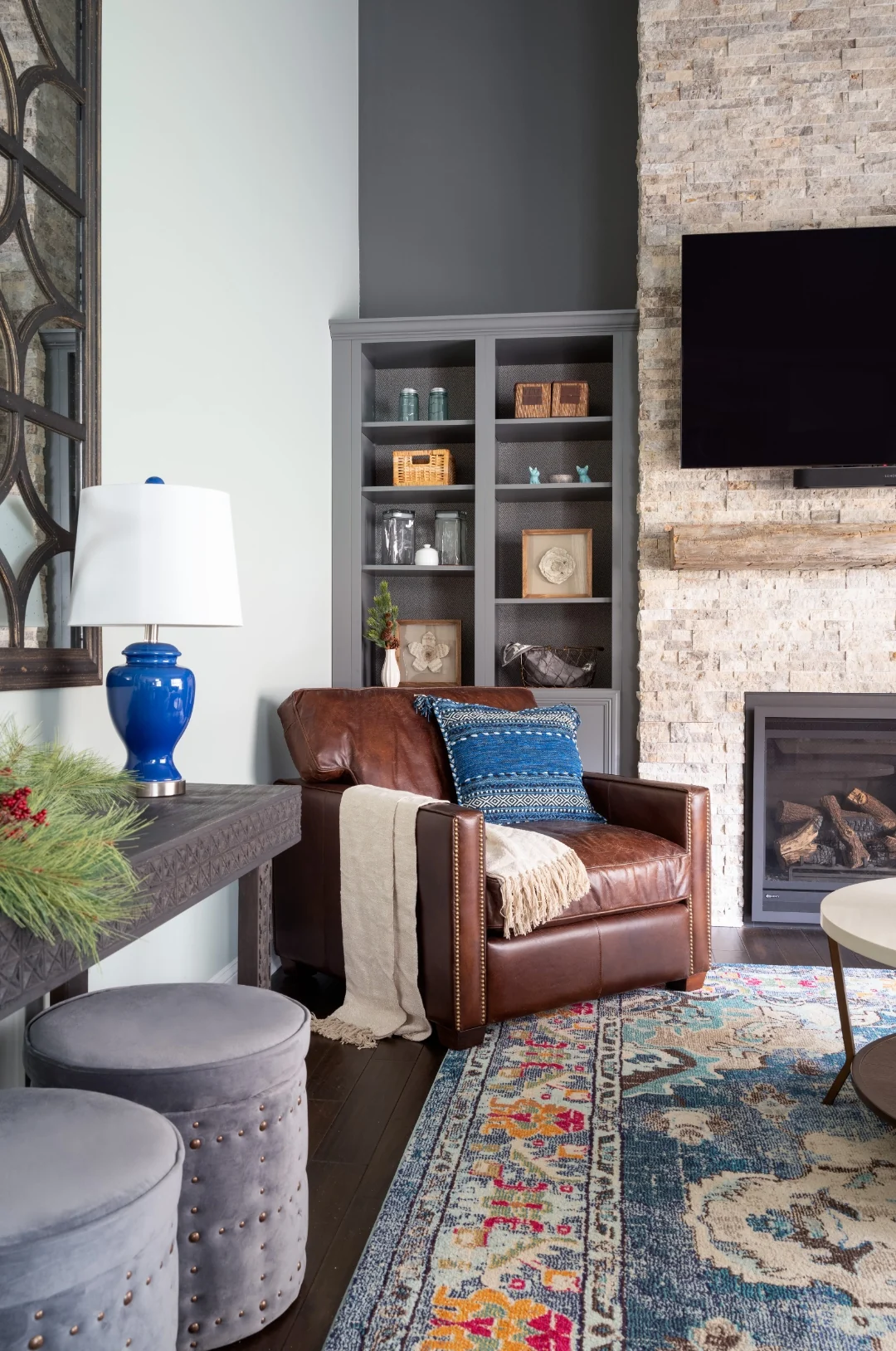 Caramel leather club chair with a woven blue lumbar pillow and ivory throw beside gray built-in bookcases and a blue ginger-jar lamp, with the stacked stone fireplace and Persian rug in the Yardley living room